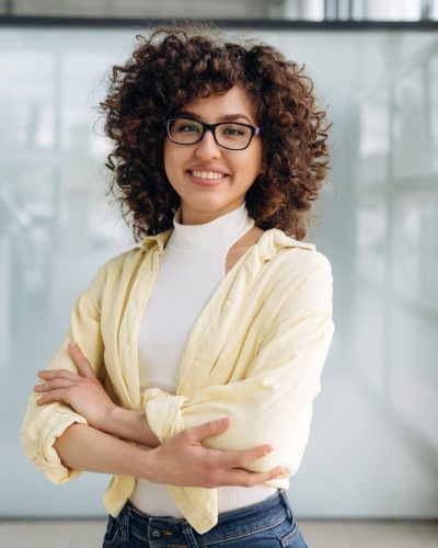 Arms crossed, standing. Beautiful woman is standing indoors against blurred wall.